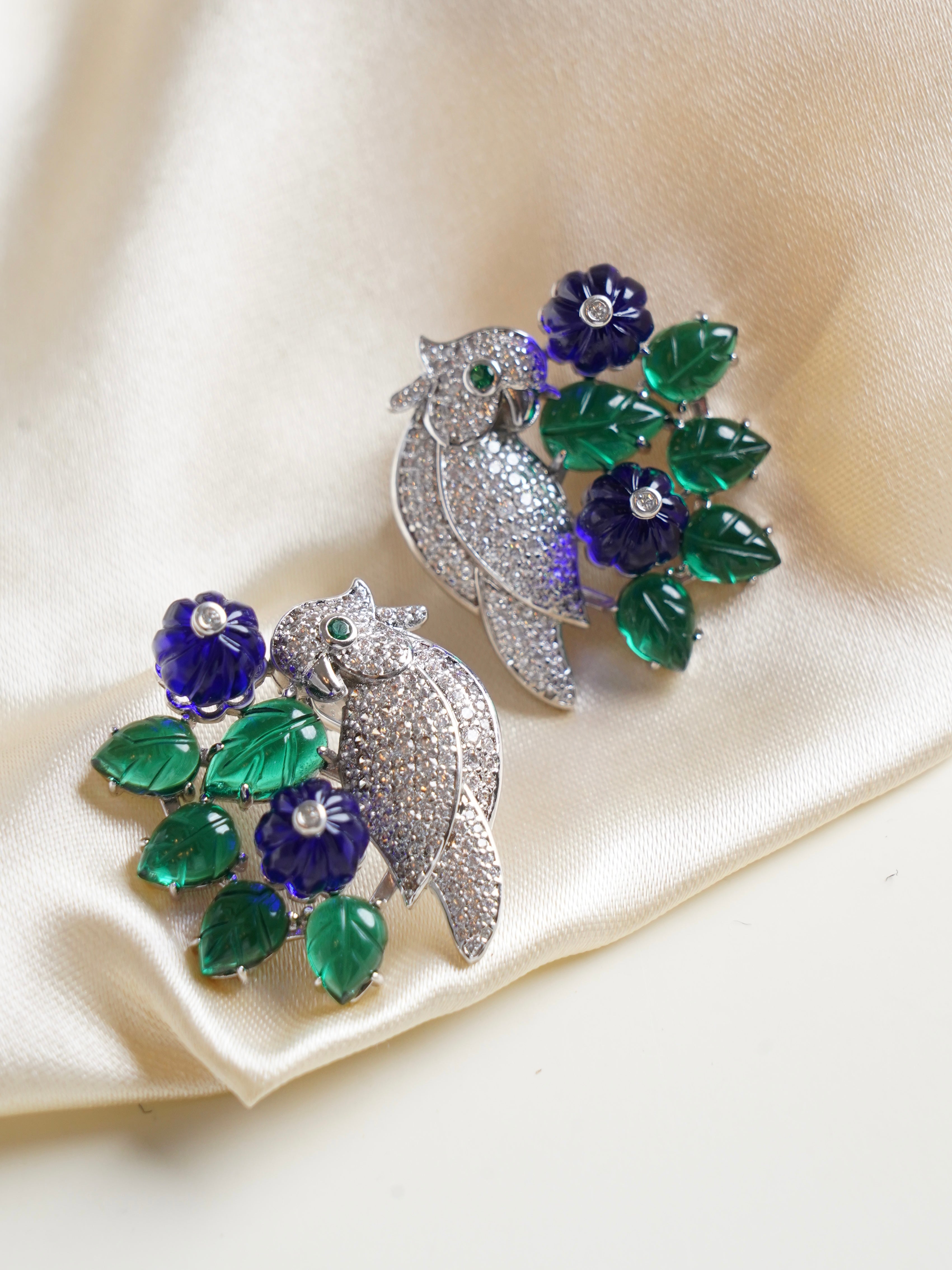 Pavé‑set bird earrings accented with carved emerald green leaves and deep violet flowers on silver base